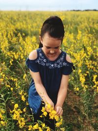 Cute girl picking yellow flower on field against sky