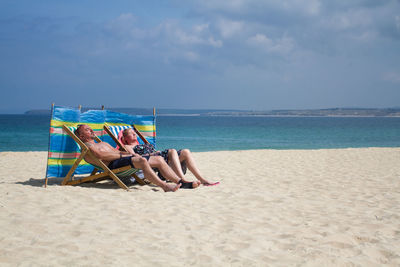 Mature couple sitting on deck chair while relaxing at beach against sky
