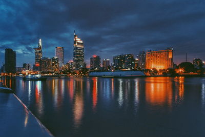 Illuminated buildings by river against sky at dusk