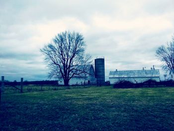 Bare trees on field by buildings against sky