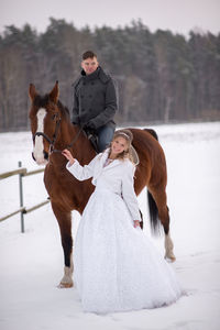 Portrait of smiling bride standing by groom sitting on horse at snow covered field