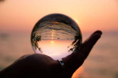 Close-up of hand holding crystal ball against sky during sunset