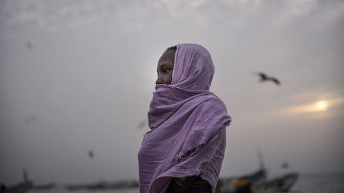 Side view of woman wearing scarf standing at beach against sky during sunset