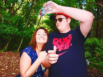 Portrait of smiling young woman holding sunglasses against trees