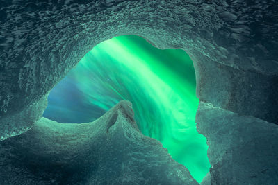 From below ice peaks surrounding hole in cave ceiling against glowing green polar lights on winter day in vatnajokull national park in iceland