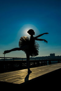 Silhouette ballerina dancing on wooden table at pier against sky during sunny day