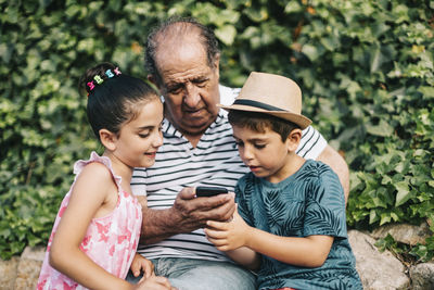 Cute boy wearing hat using smart phone while sitting with grandfather outdoors