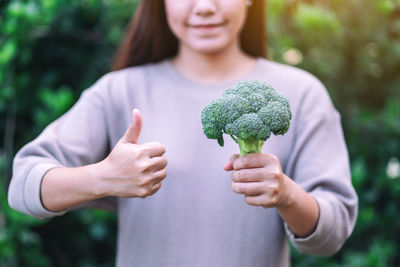 Midsection of woman holding broccoli while showing thumbs up outdoors