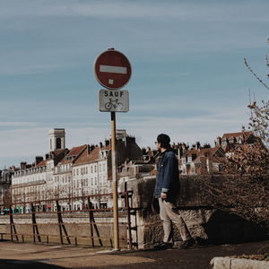 Side view of young man standing on footpath against sky in city