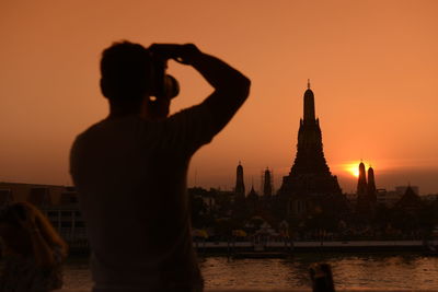 Rear view of man photographing wat arun temple against sky during sunset