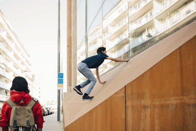 Carefree teenage boy climbing on wall while friend standing at sidewalk in city