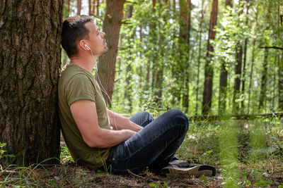 Side view of young man sitting on tree trunk in forest