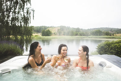 Cheerful female friends enjoying wine in hot tub against lake during weekend getaway