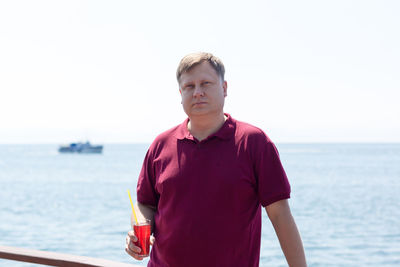 An adult blond man in a bright burgundy t-shirt with a glass of strawberry juice is standing