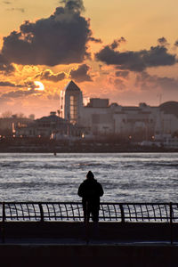 Rear view of silhouette man standing by railing against river during sunset