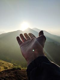 Cropped hand of man gesturing towards mountain against sky during sunrise