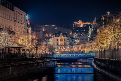 Illuminated bridge over river by buildings in city at night