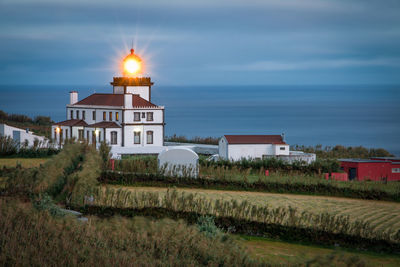 Illuminated lighthouse by sea against cloudy sky