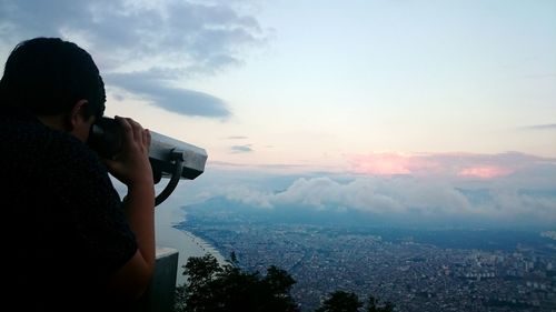 Side view of boy looking through coin-operated binoculars against sky during sunset