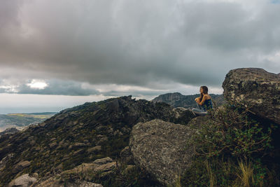 Man sitting on rock looking at mountain against sky