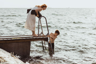 Side view of woman guiding while son moving down on ladder in sea