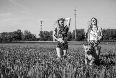 Female friends with dog walking on grassy field against sky