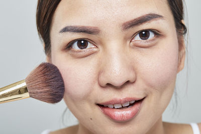 Close-up portrait of beautiful mid adult woman applying make-up over gray background