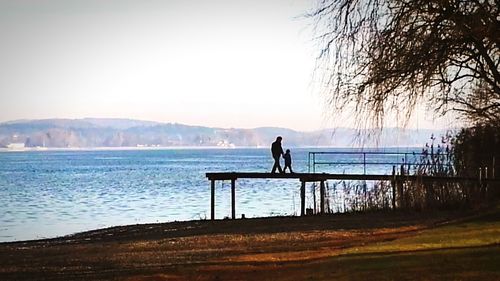 Rear view of silhouette man on bench in front of lake against clear sky