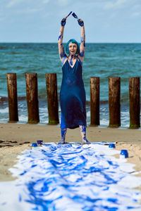 Woman performance artist smeared with blue gouache paints happily dancing hands up on beach