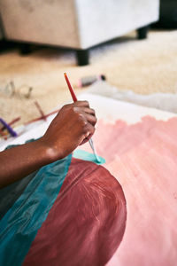 From above of crop anonymous black female with paintbrush drawing colorful picture on paper while sitting on floor at home