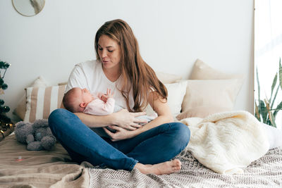 A tender caring mother lulls a newborn baby in her arms, sitting on the bed.