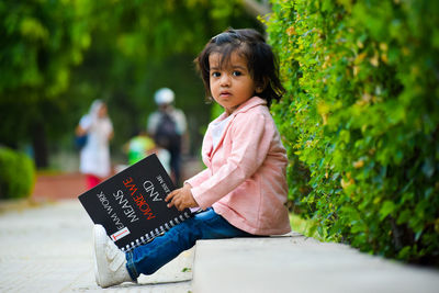 Portrait of girl holding book while sitting by plant at park
