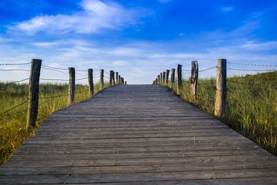 Wooden boardwalk leading towards plants against cloudy sky