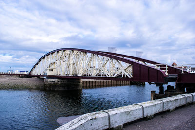 Metallic bridge over river against cloudy sky