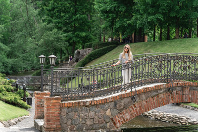 A woman stands on an old stone bridge with a forged fence in the park in the summer