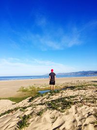 Full length rear view of man standing at beach against blue sky