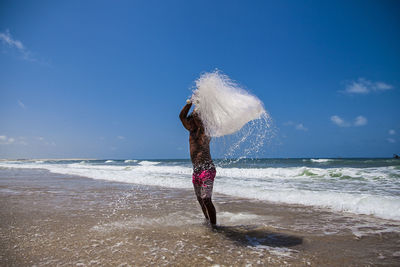 Shirtless fisherman holding fishing net while standing on shore at beach against sky