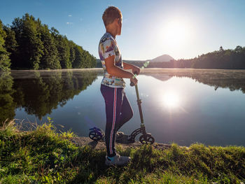 Blond hair boy with scooter stands on a park path along lake. childhood sports concept. evening sun