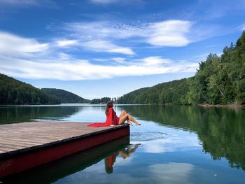 Woman in red dress sitting on a wooden pontoon near the lake on a beautiful sunny day