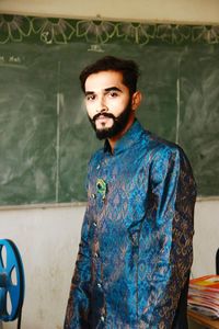 Portrait of young man in traditional clothing standing against green board in classroom