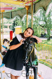 Beautiful brunette girl in a black dress poses on a bright carousel with horses