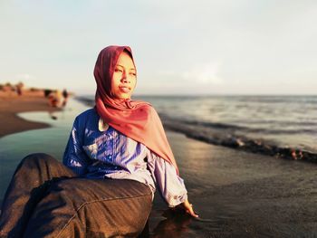 Young woman sitting at beach against sky