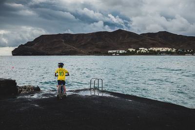 Rear view of boy on bicycle by sea against cloudy sky