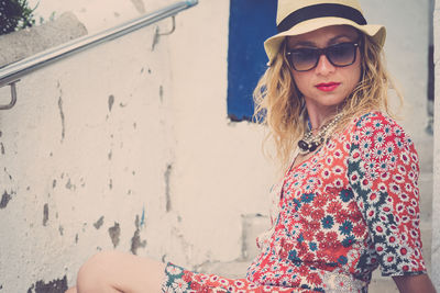 Portrait of woman wearing sunglasses sitting against wall