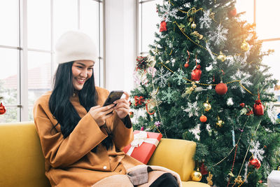 Young woman using mobile phone while sitting on sofa by christmas tree