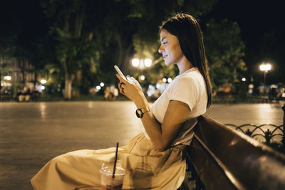 Woman using smart phone sitting on park bench at night