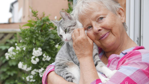 Portrait of senior woman cuddling tabby cat by house