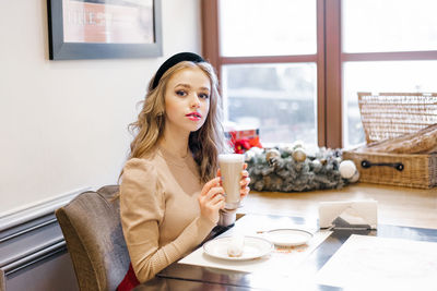 A beautiful young girl is sitting at a wooden table by the window and drinking coffee in a cafe 