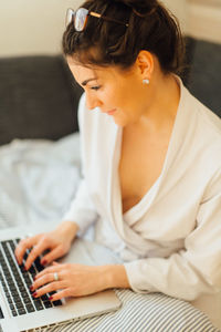 High angle view of woman in bathrobe using laptop while sitting on bed at home