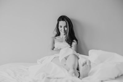 Portrait of smiling woman with blanket sitting on bed against wall